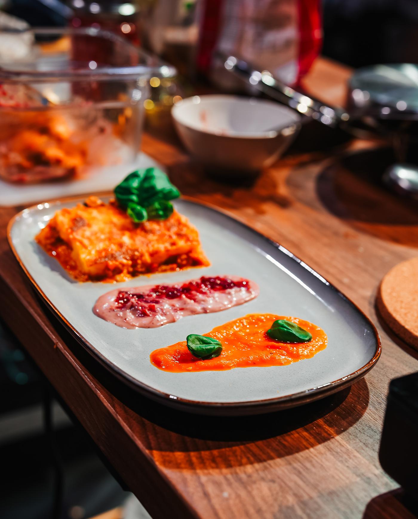 An oval plate on a wooden table features a piece of lasagna with a savory portion, complemented by colourful sauces and garnished with herbs. Surrounding utensils and ingredients add to the culinary presentation.