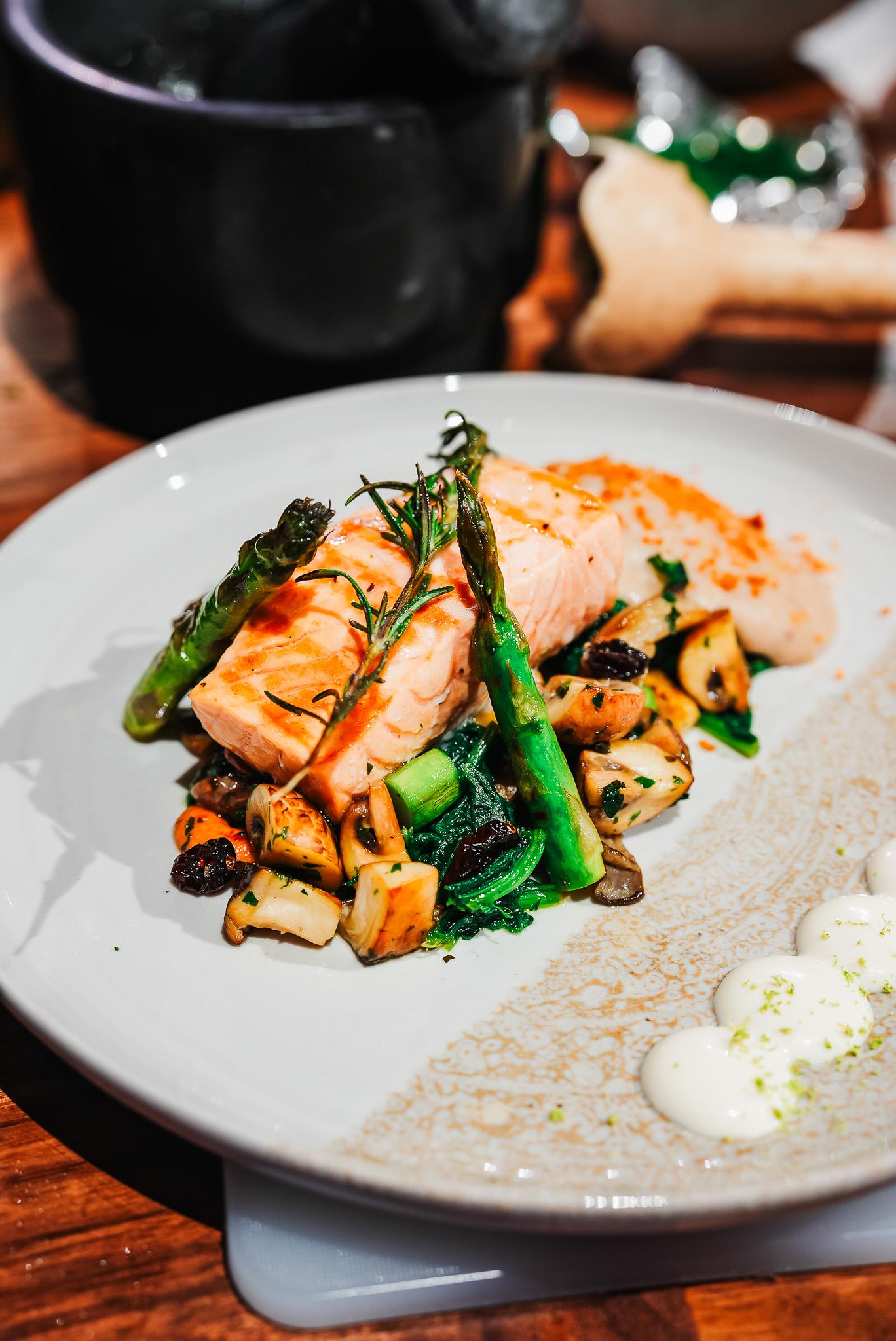 A plate featuring grilled salmon, asparagus, and sautéed vegetables, elegantly arranged with a creamy sauce on the side. The dish is set on a wooden table, accompanied by cooking utensils and ingredients.