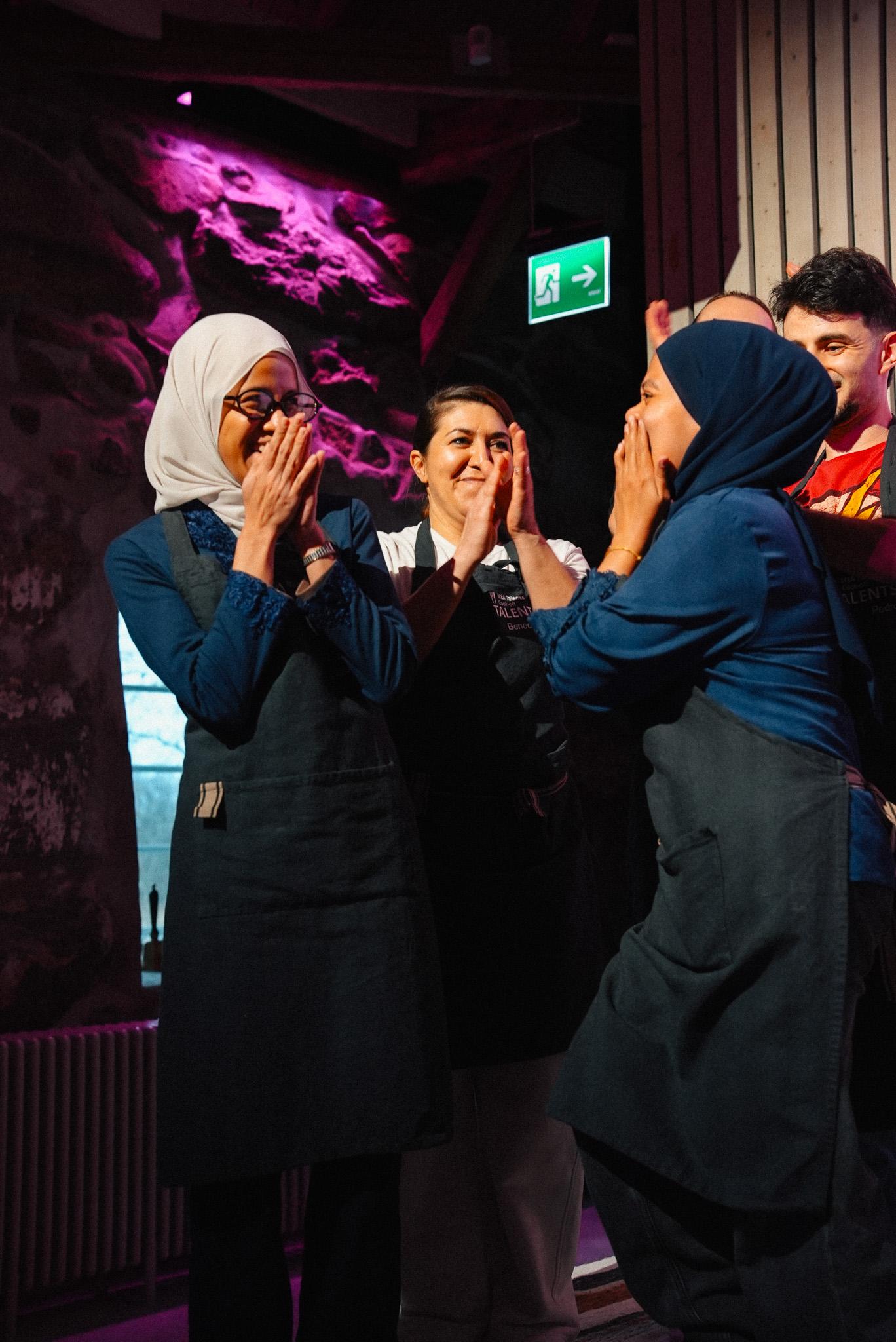 Two people wearing dark aprons and hijabs share a moment of surprise and excitement in a dimly lit room with vibrant purple lighting in the background.