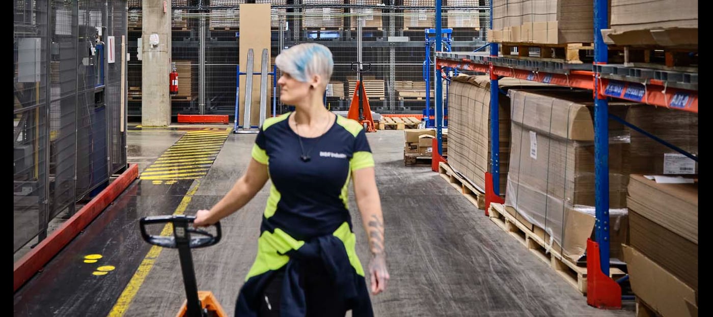 A person in a warehouse setting, wearing safety gear and pulling a pallet jack.