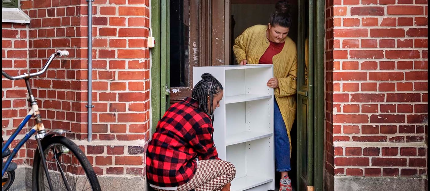 Two people stand in the doorway of an apartment building. They are moving a white shelf unit through the doorway.