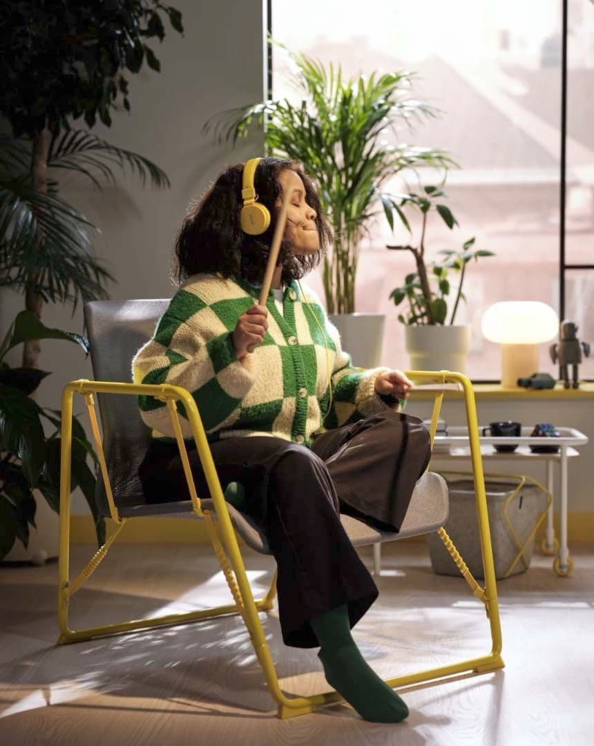A girl sits on a yellow and grey easy chair from the BRÄNNBOLL collection. She is listening to music and playing pretend drums with drumsticks.