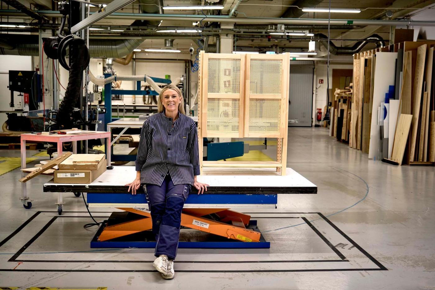 Designer Ebba Strandmark leans against a table in a workshop in front of wooden storage furniture.