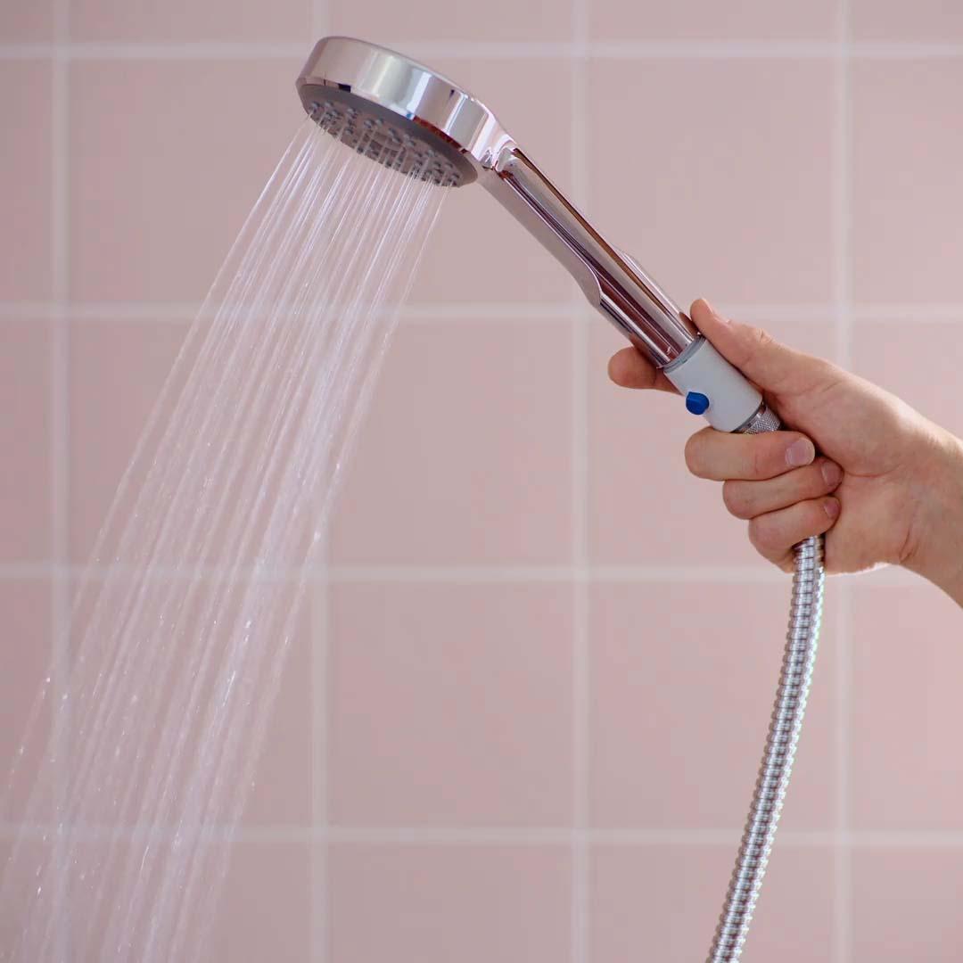 A person holding a hand shower with a blue booster button in a bathroom with pink tiles.
