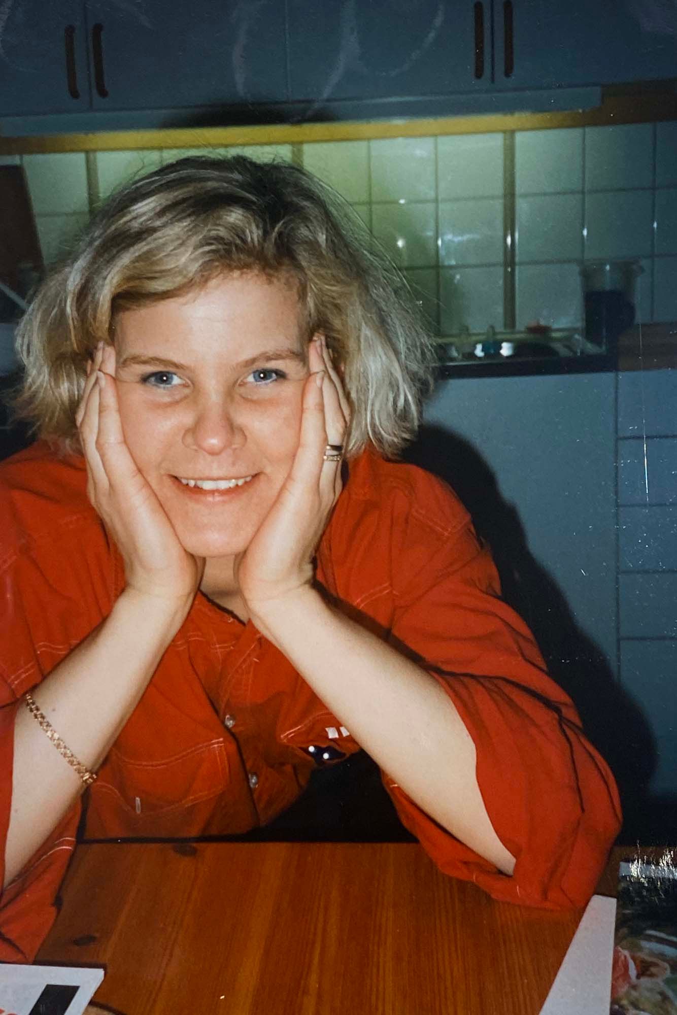 A vintage image of Fillipa sitting in her kitchen wearing a red IKEA uniform. She sits with her hands in her face, smiling.