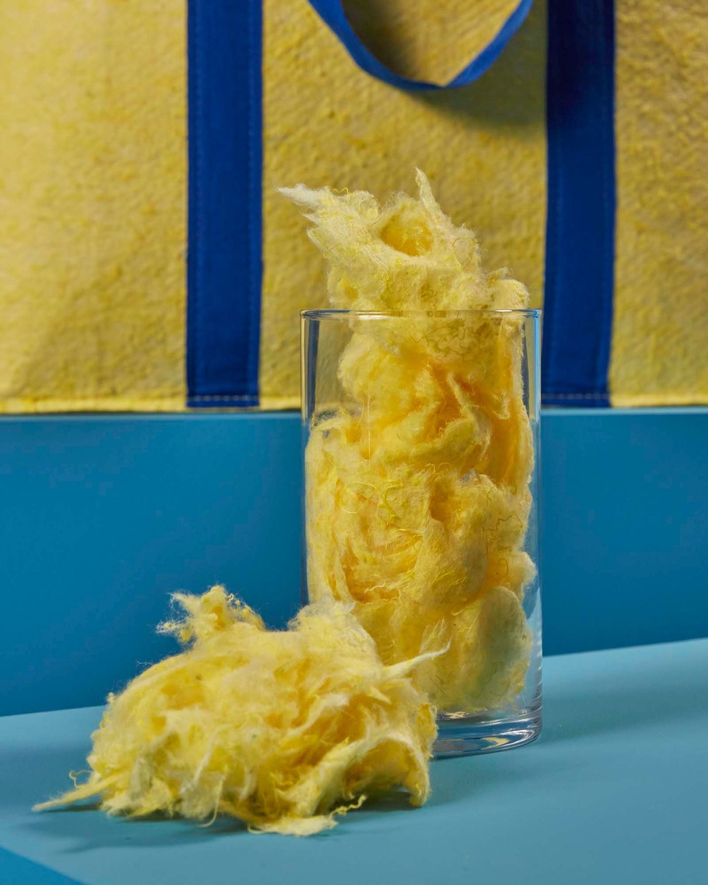 A cylindrical glass container filled with shredded yellow fibres, with a bag from the VÄXELBRUK collection in the background.