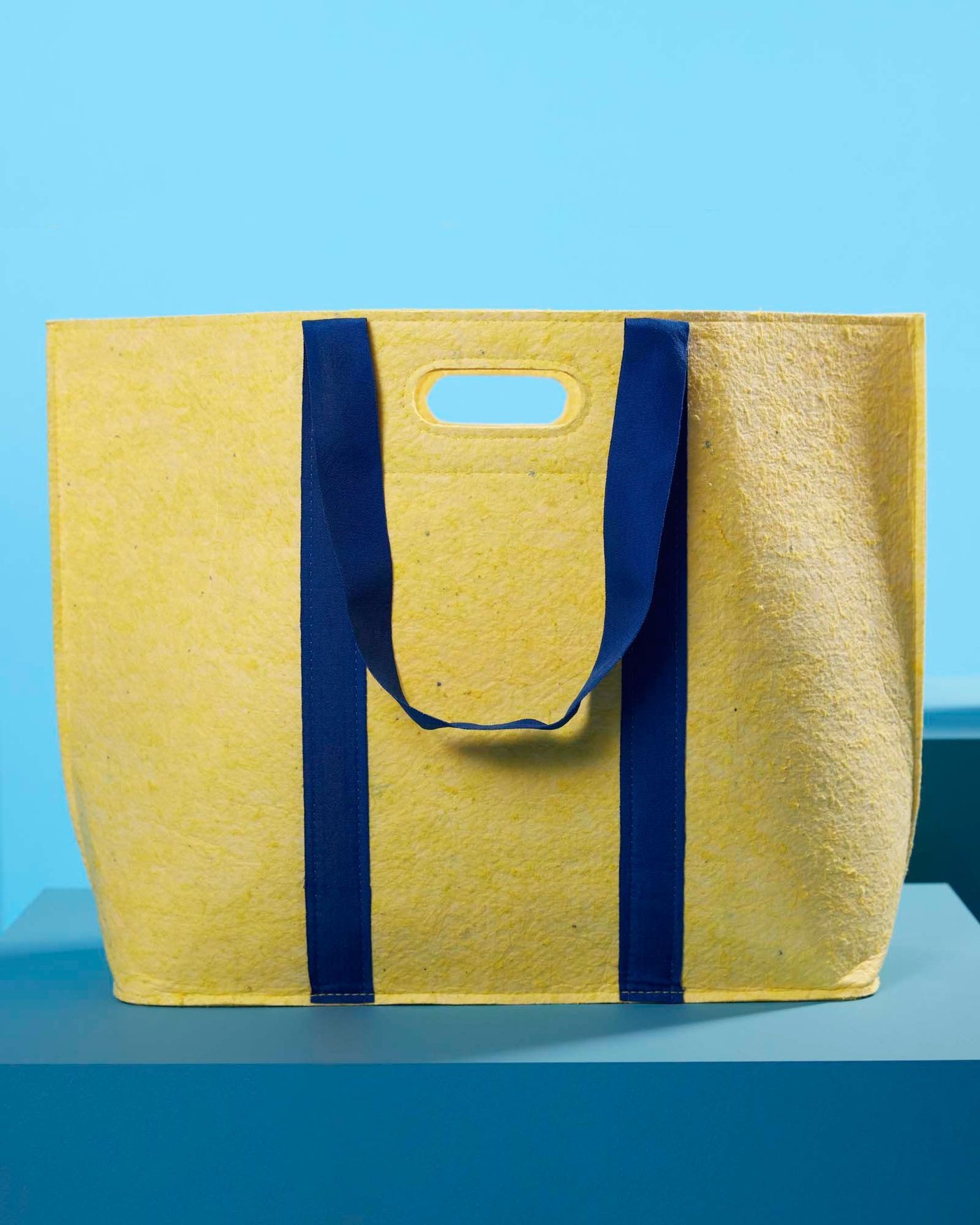 Yellow felt bag from the VÄXELBRUK collection displayed on a blue plinth.