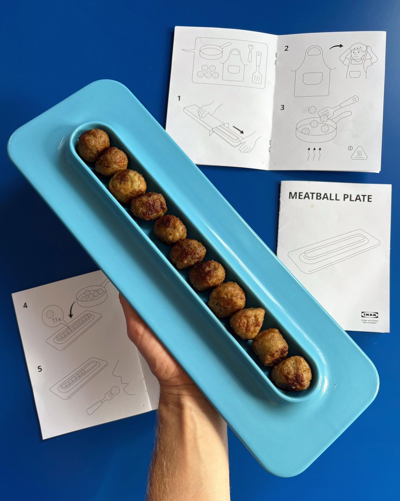 A hand holds a blue plate filled with meatballs lined in a row, against a blue surface and instruction manuals.