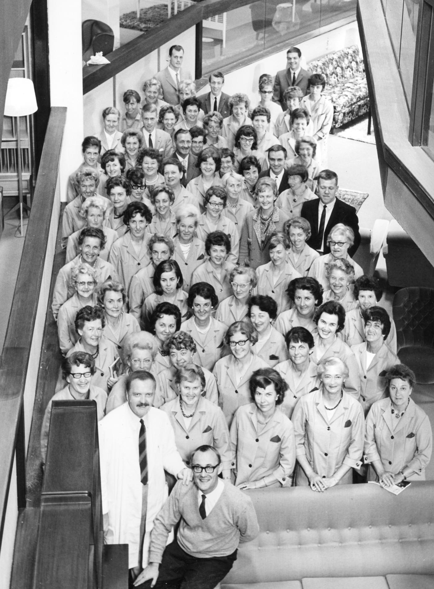 A black-and-white image of Ingvar Kamprad sitting on the arm of a sofa with many IKEA co-workers behind him.