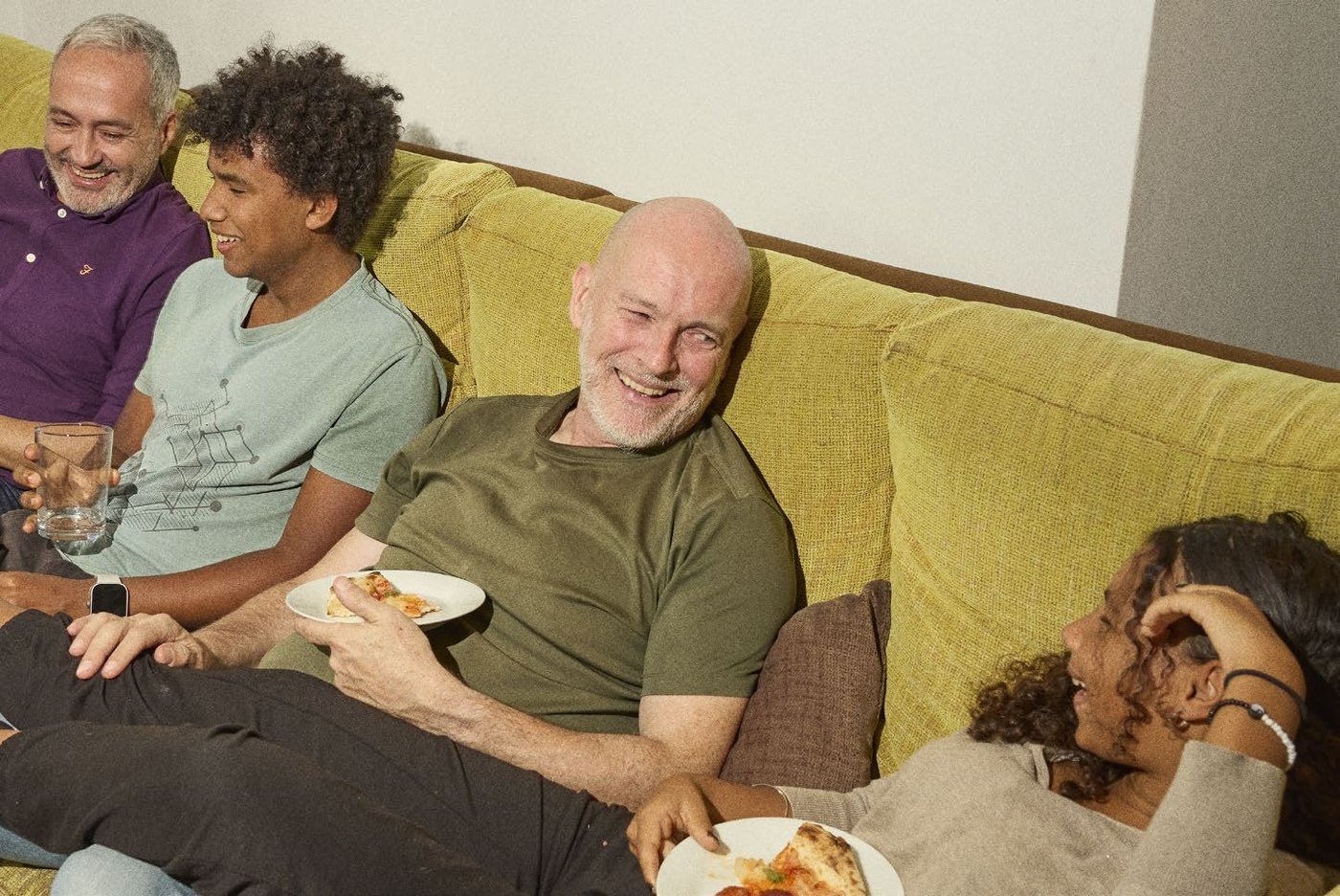 A family sit on a long green sofa, while eating pizza on their laps. They are talking and smiling.