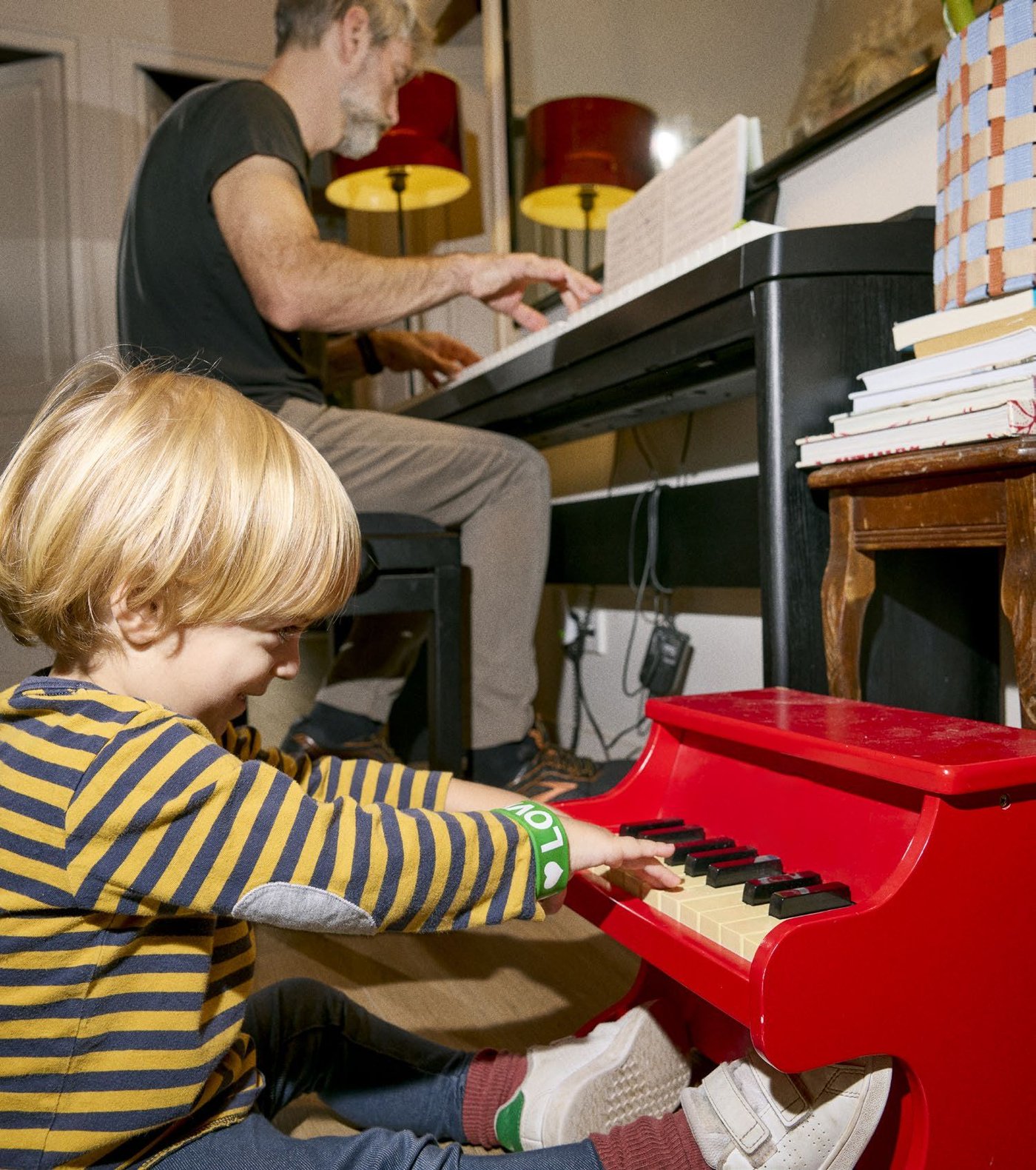 David plays a piano next to his child, who is playing a small, red children's piano.
