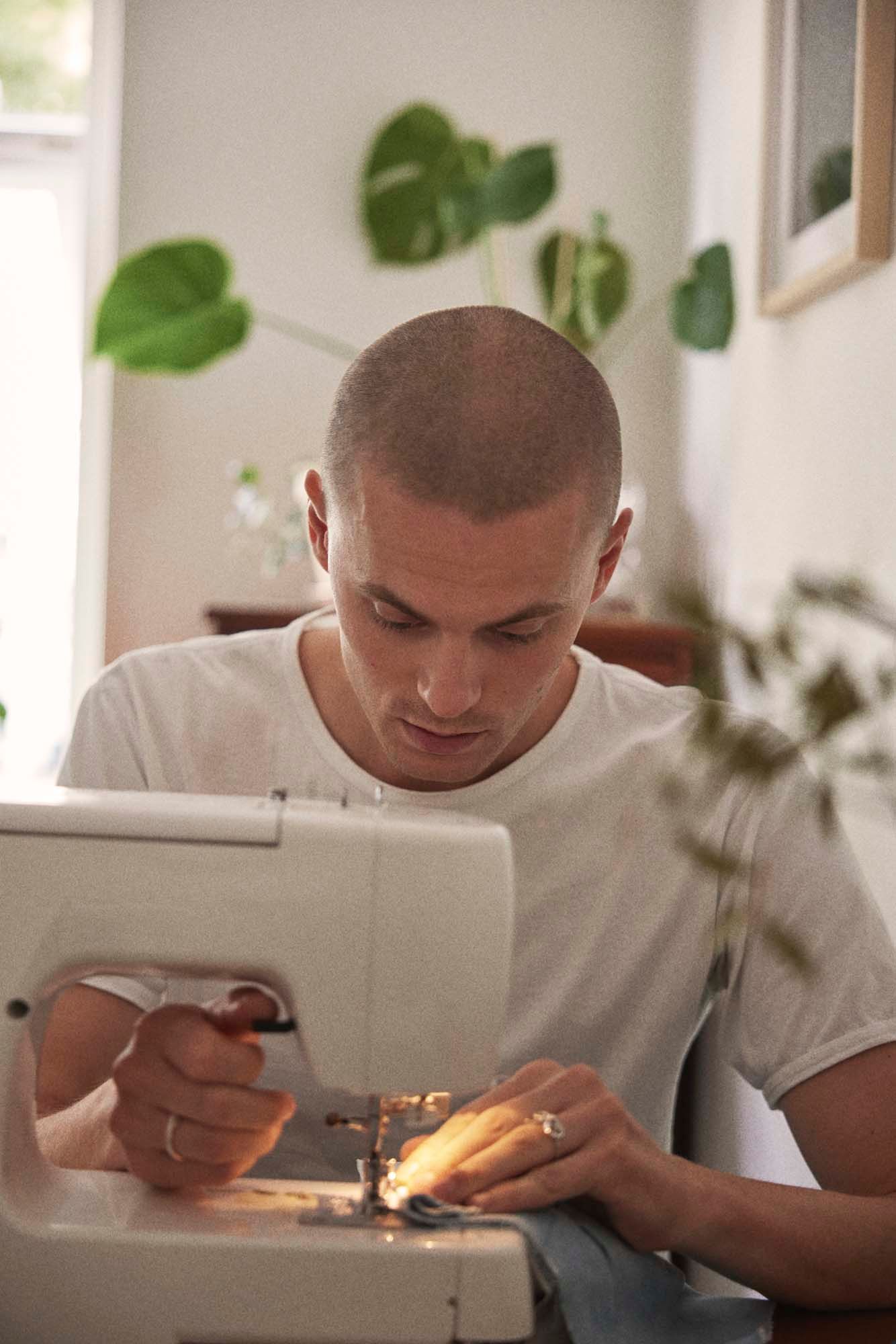 A young man in a white tshirt uses a sewing machine.
