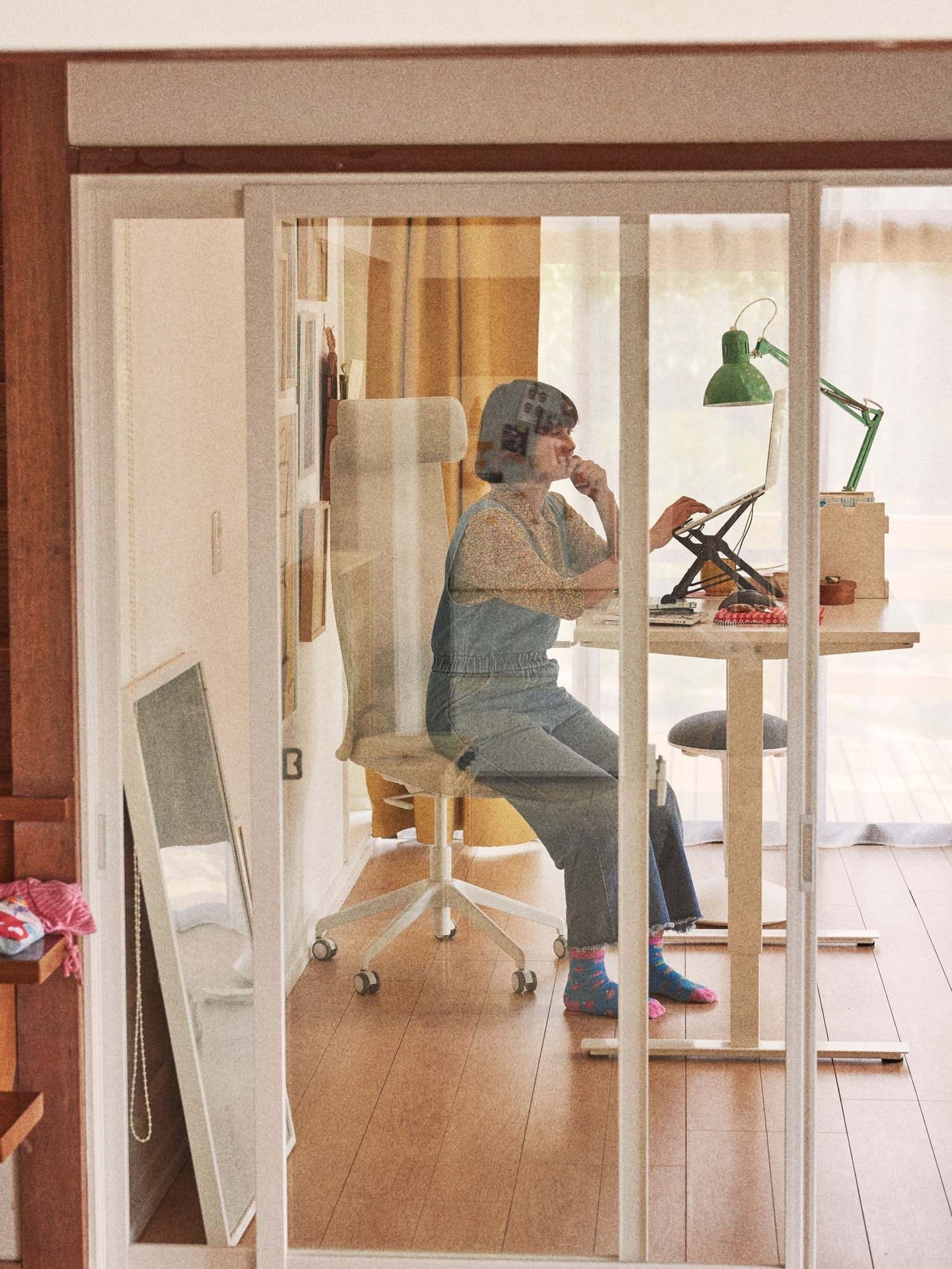 A person wearing overalls sits on a stool at a desk in a home workspace.