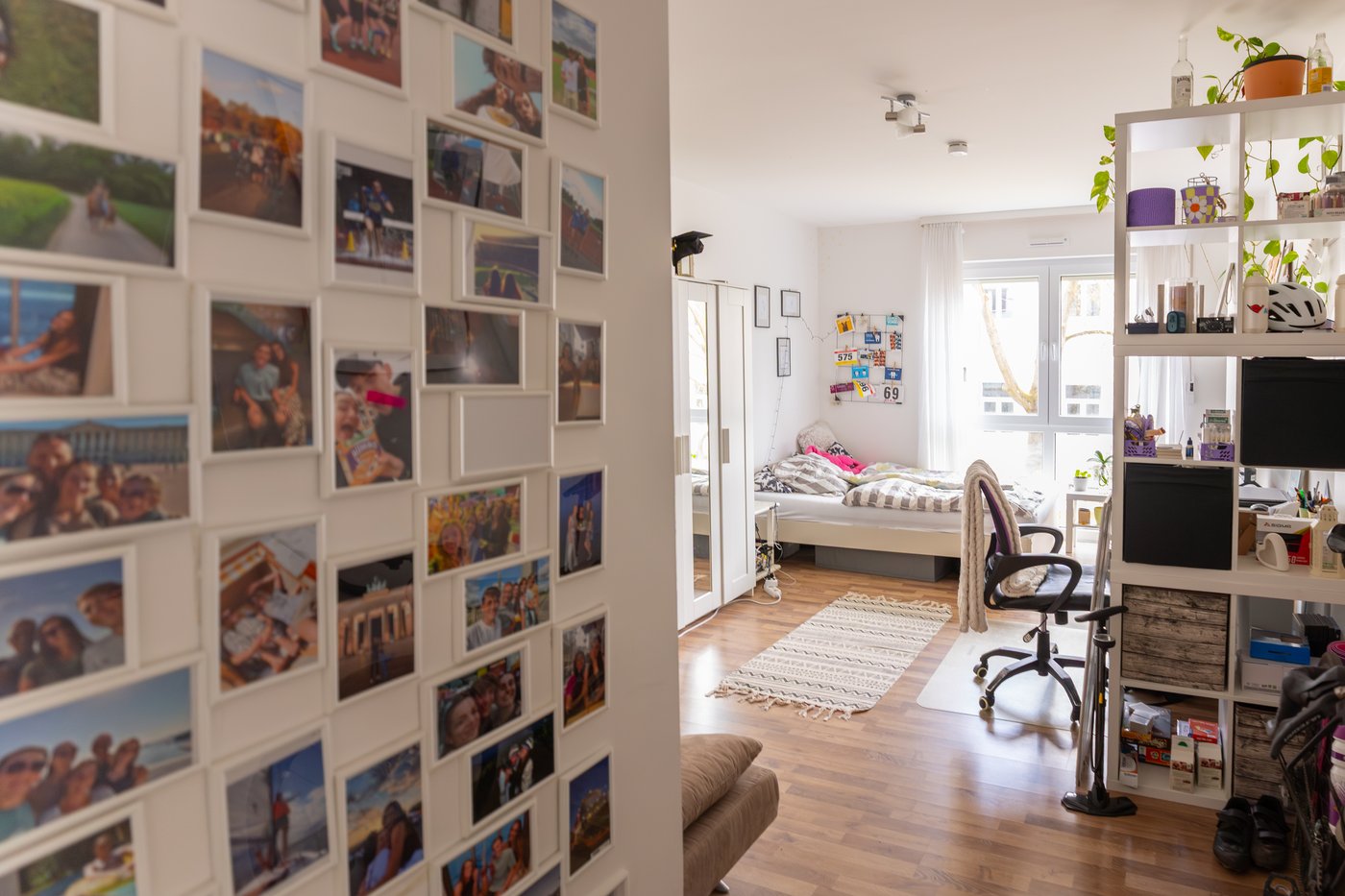 A view into a room with white walls, with a gallery wall filled with photographs, open shelving with a desk behind and a bed.