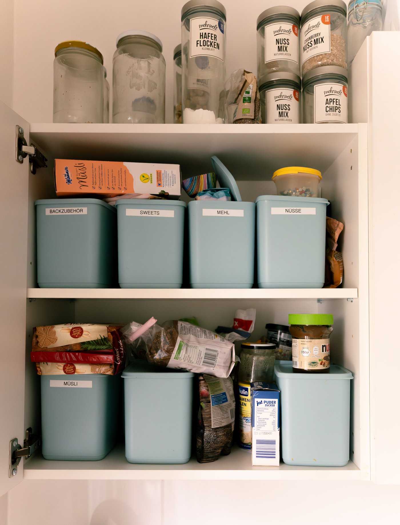 An array of food items surround neatly arranged light-blue storage containers with labels inside a white kitchen cupboard.