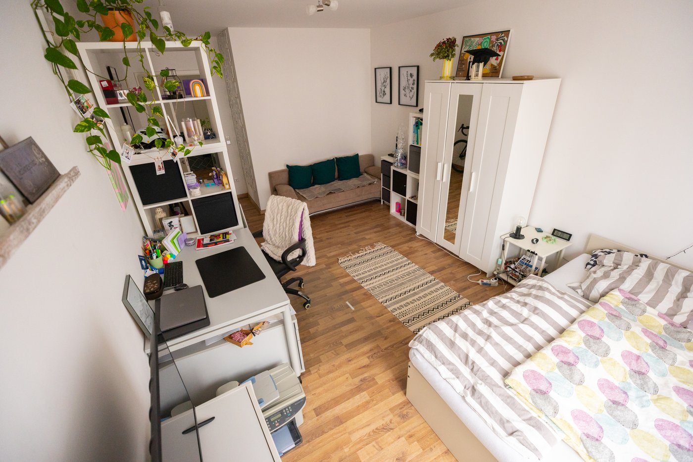 An angled view of a bed, desk, wardrobe, open shelving and sofa in a white studio apartment with a wood floor.