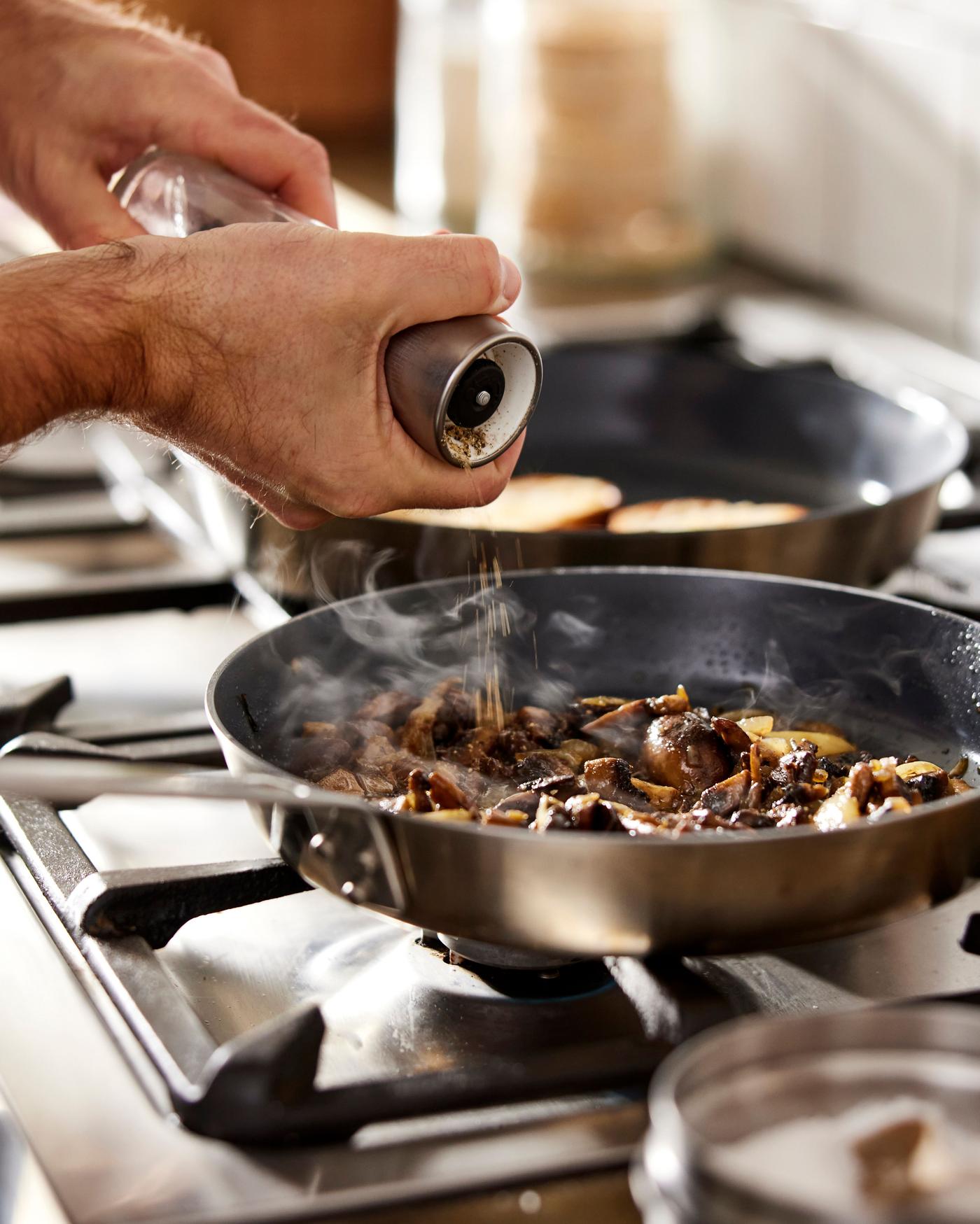 The HEMKOMST pan on a stove with the fire on. In the pan different vegetables are being cooked. Someone is holding a peppermill above the pan, sprinkling some pepper onto the vegetables.