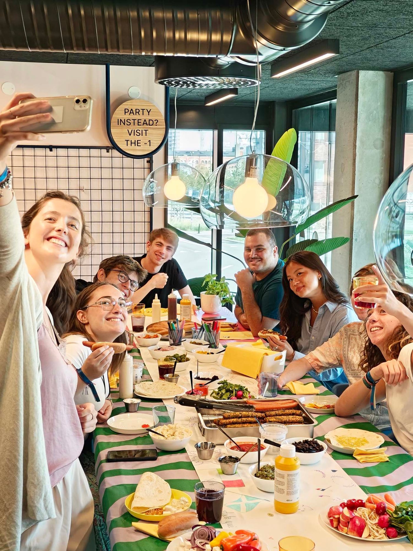 A group of students sit around a table with food including hot dogs. One person takes a selfie.
