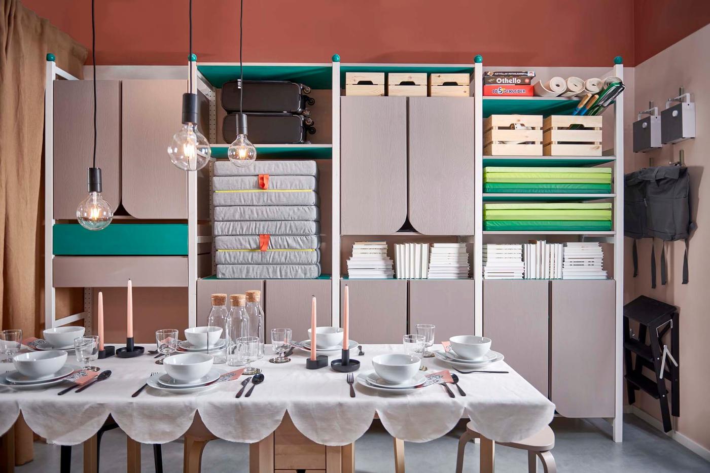 A room with grey storage filled with boxes and cushions etc., in the background and a set table with hanging lamps in the foreground.