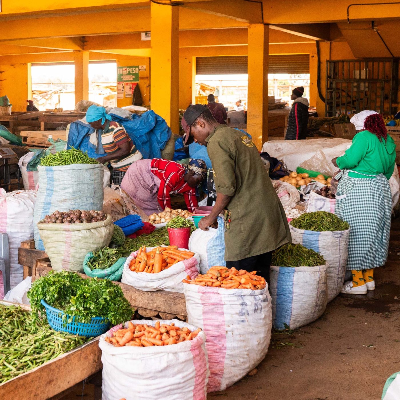 People walk between bags of fruit and vegetables in a market.