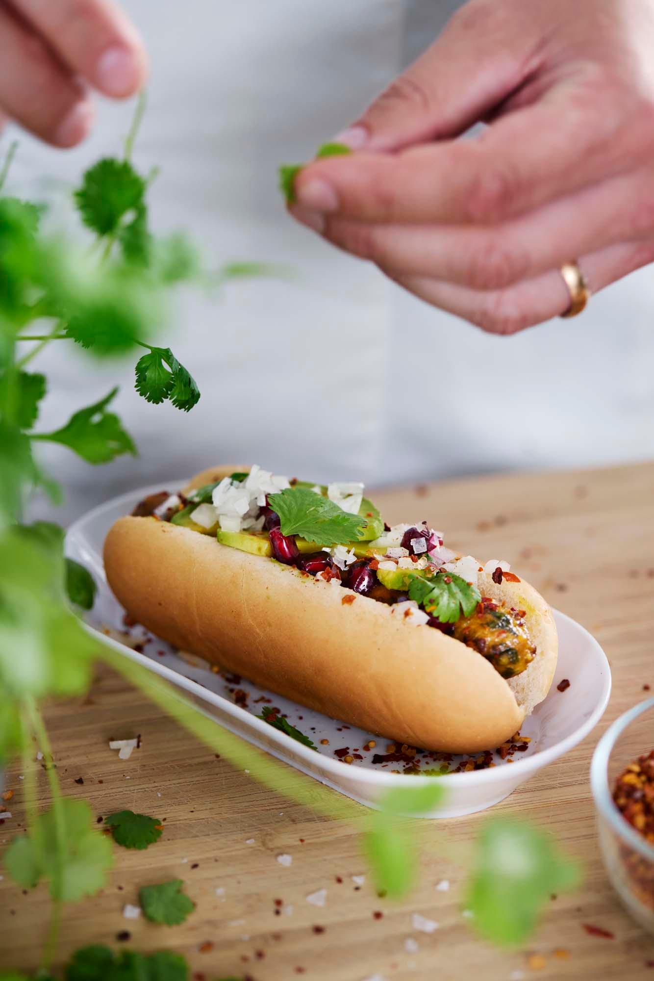 Chef's hands putting the final garnish on a vegetable sausage in a hot dog bun. 