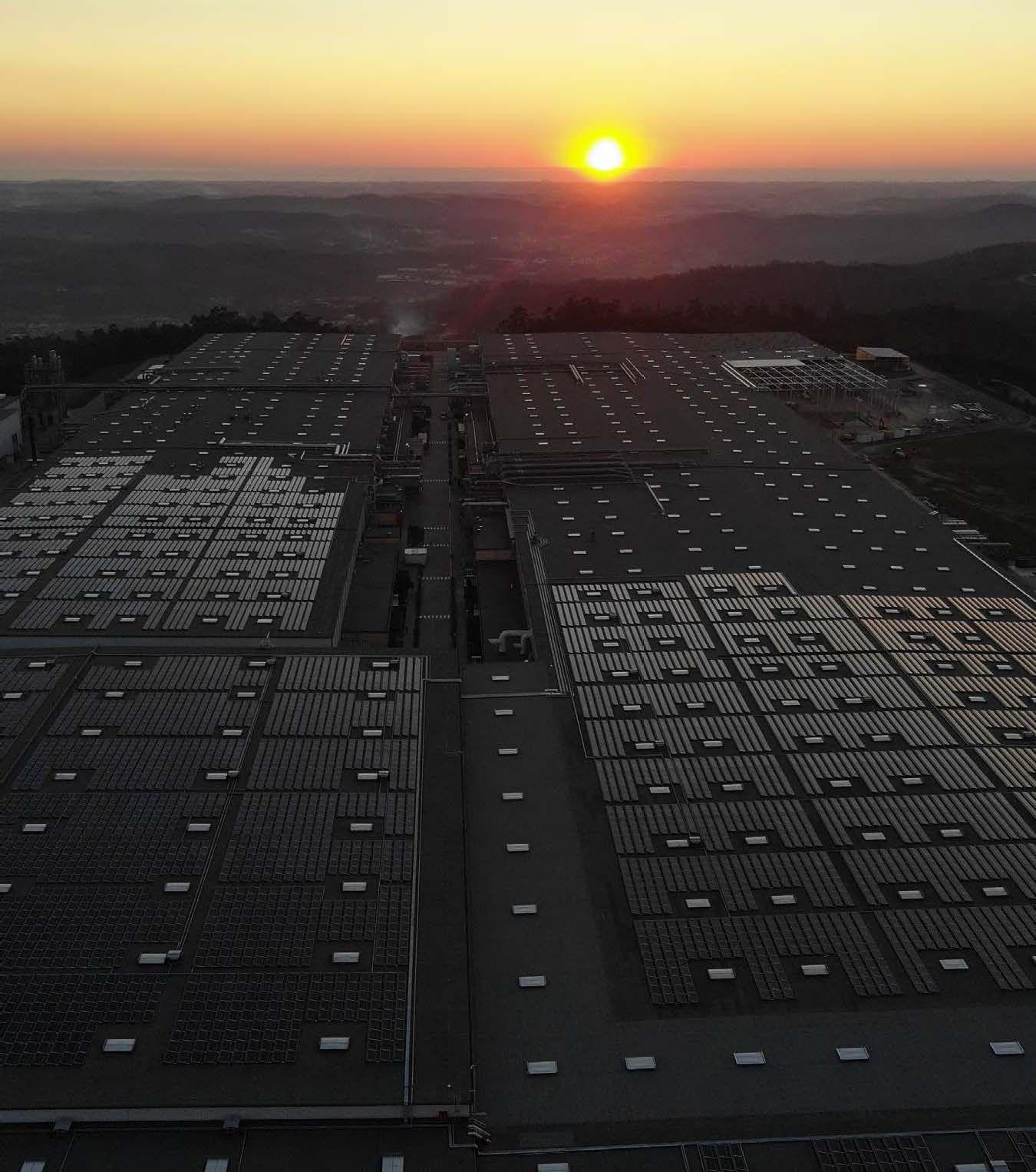 Darkness falls as the sun sets over the land. In the foreground are many solar panels. In the background are rural and urban landscapes.