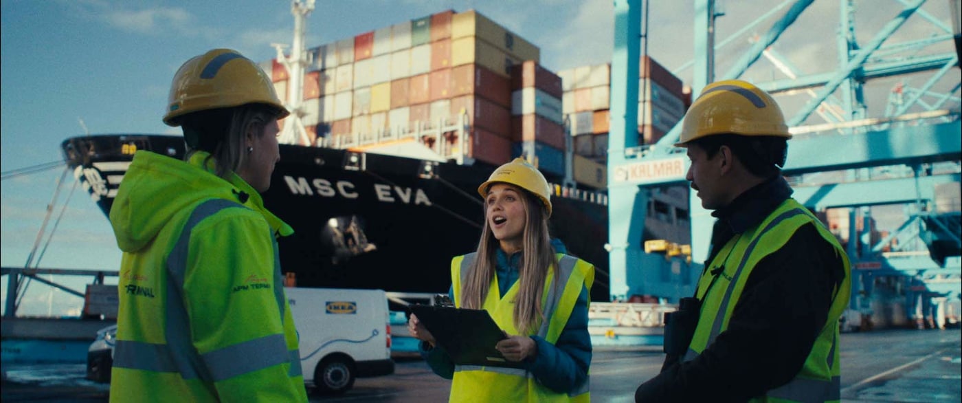Sabine and two other people stand talking at a port, dressed in helmets and reflective vests. A ship loaded with containers is visible behind them.