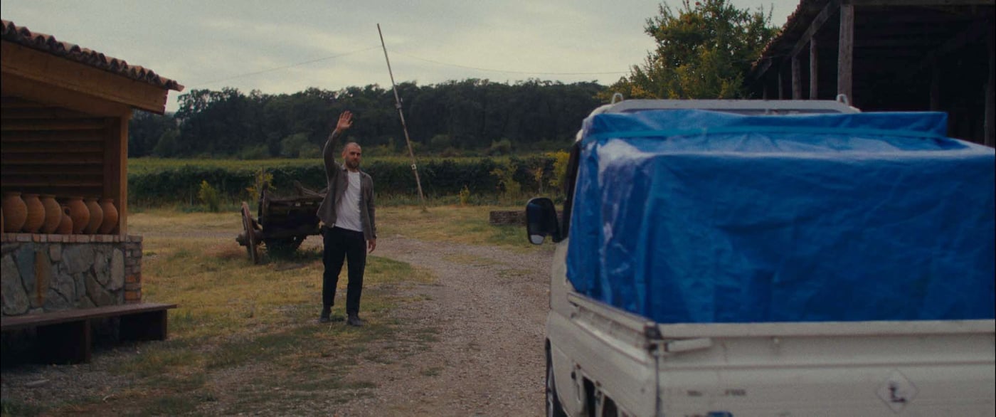 A man waves to an approaching delivery truck, with a blue tarpaulin covering the load. 