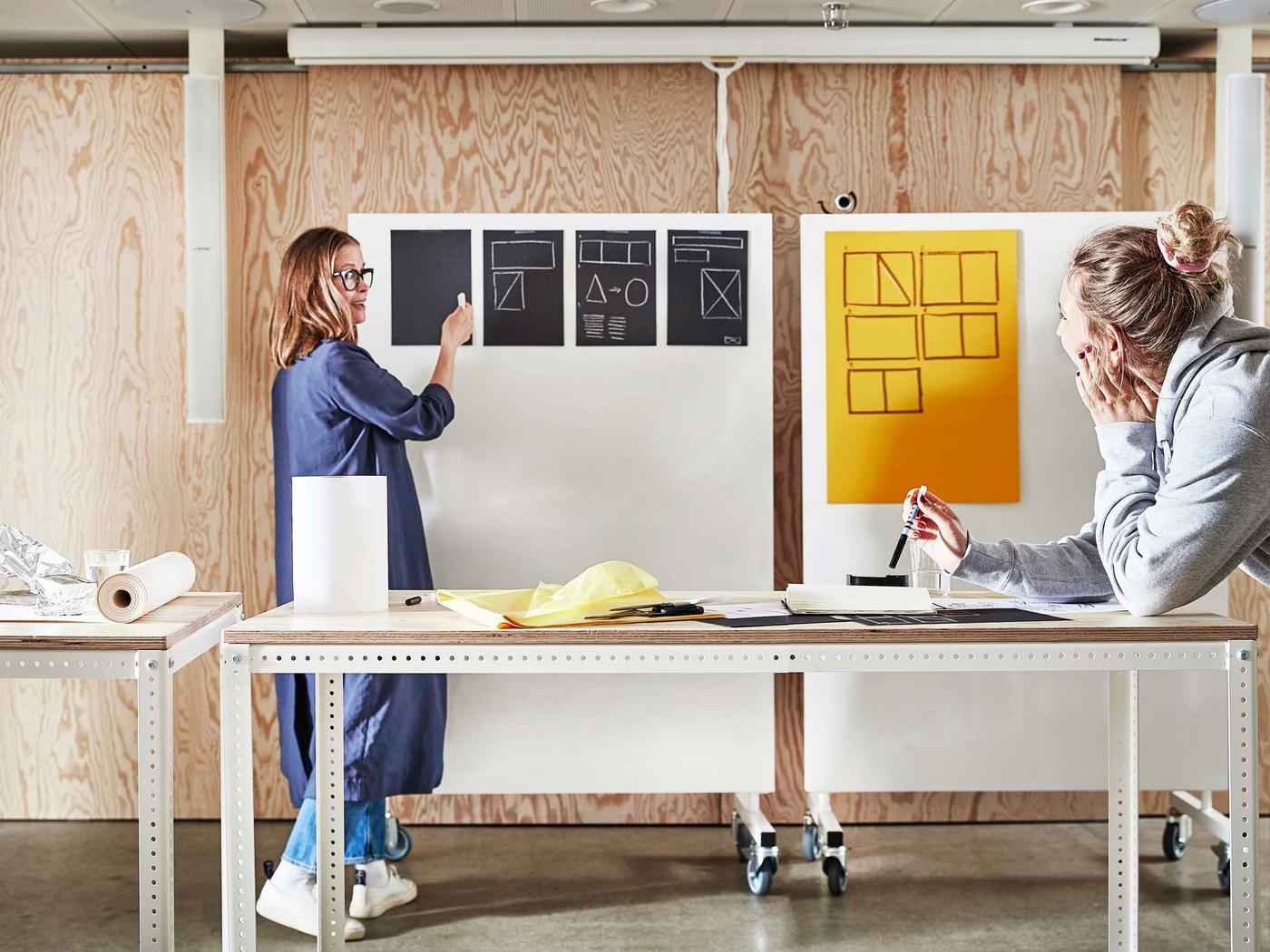 A person writes and explains on a display board while a colleague watches while leaning on a table.