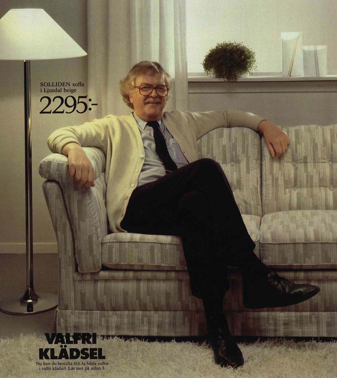 A man sits on a patterned sofa smiling. He sits near a lit floor lamp.