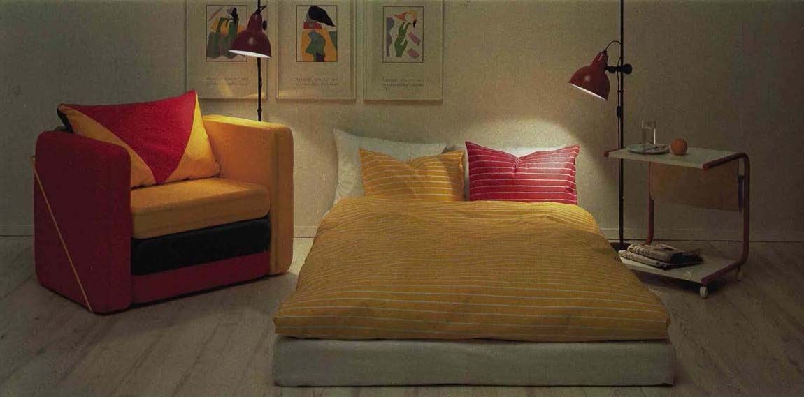 A darkened living room with a red and yellow chair beside an open sofa bed with a yellow duvet. Floorlamps shine a spotlight on the chair and bed.