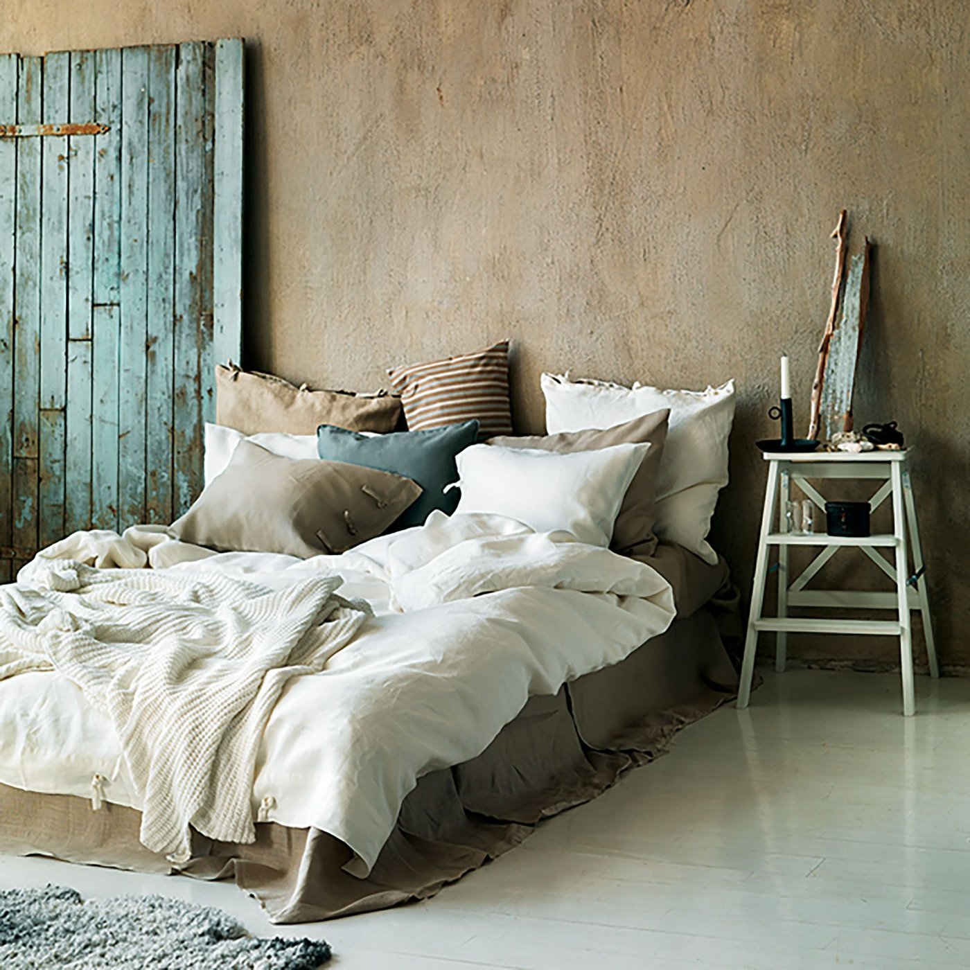 The 2013 IKEA catalogue featuring a bed covered with throws and cushions in muted natural colours. To the left is a blue rough looking wood door and to the right, a ladder doubles as a bedside table.