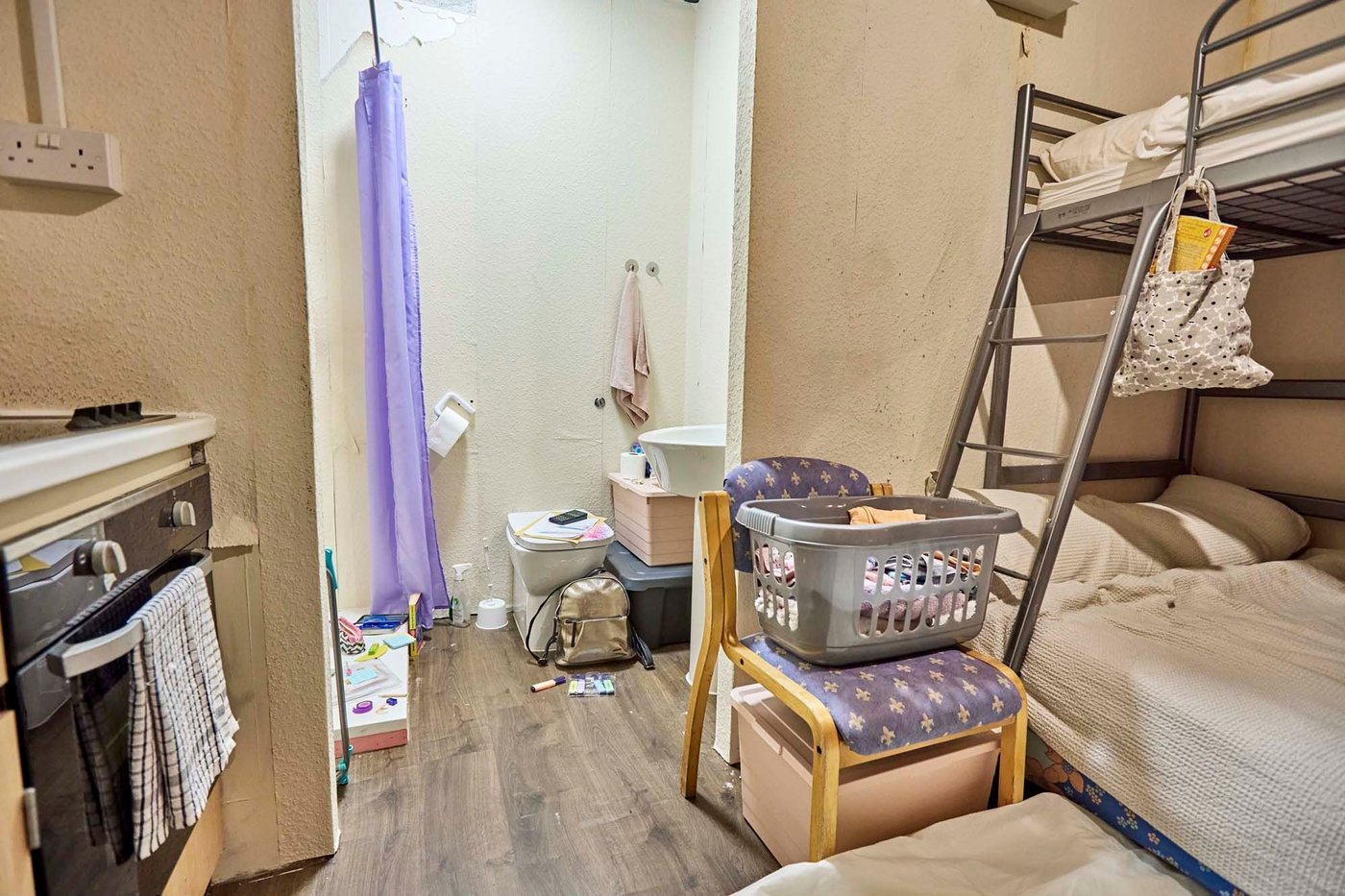 A small, harshly lit roomset with a bunk bed, extra mattress, tiny kitchenette and bathroom.