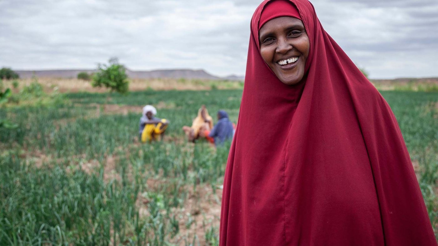 A woman in a red chador stands in a field, smiling at the camera.
