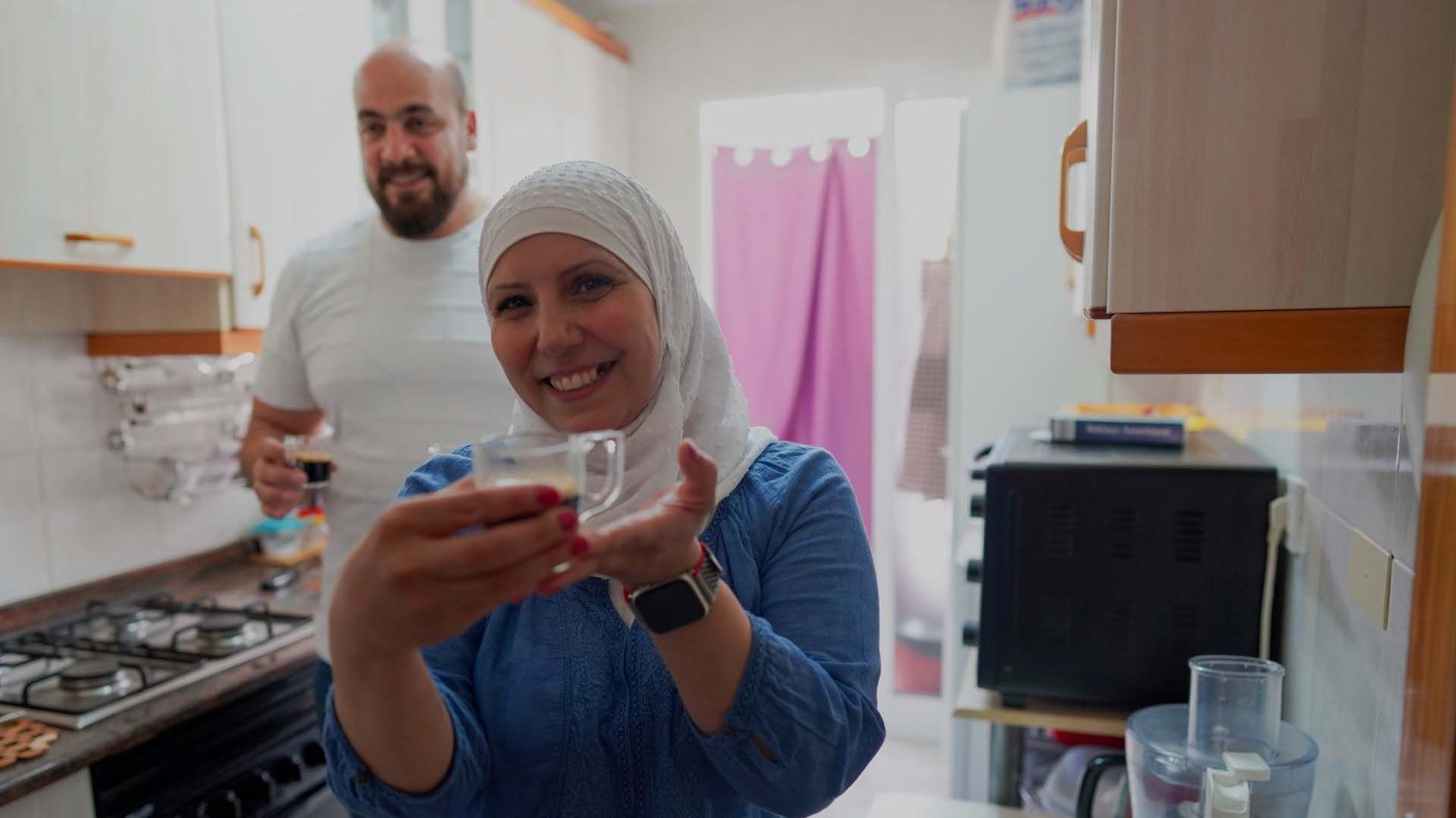 A man and woman stand in their kitchen smiling. She offers a cup of tea.