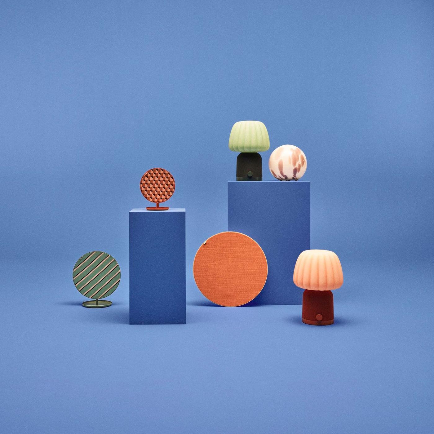 A group of colourful lamps and speakers designed by Teklan, against a blue background. Two lamps and a speaker stand on blue podiums.