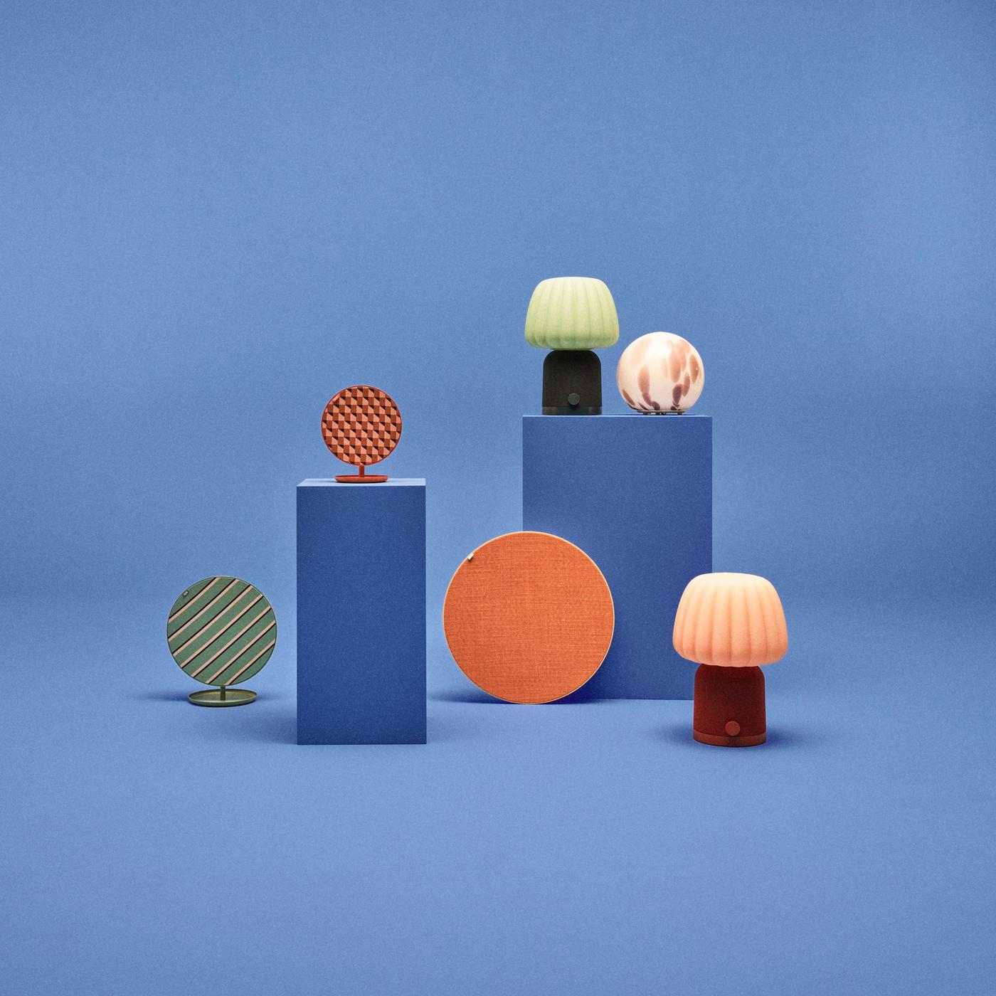 Two table lamp-shaped KULGLASS speakers and three round SOLSKYDD speakers in different sizes, colours and patterns, alongside a spherical FADO lamp. All are set against a blue background.