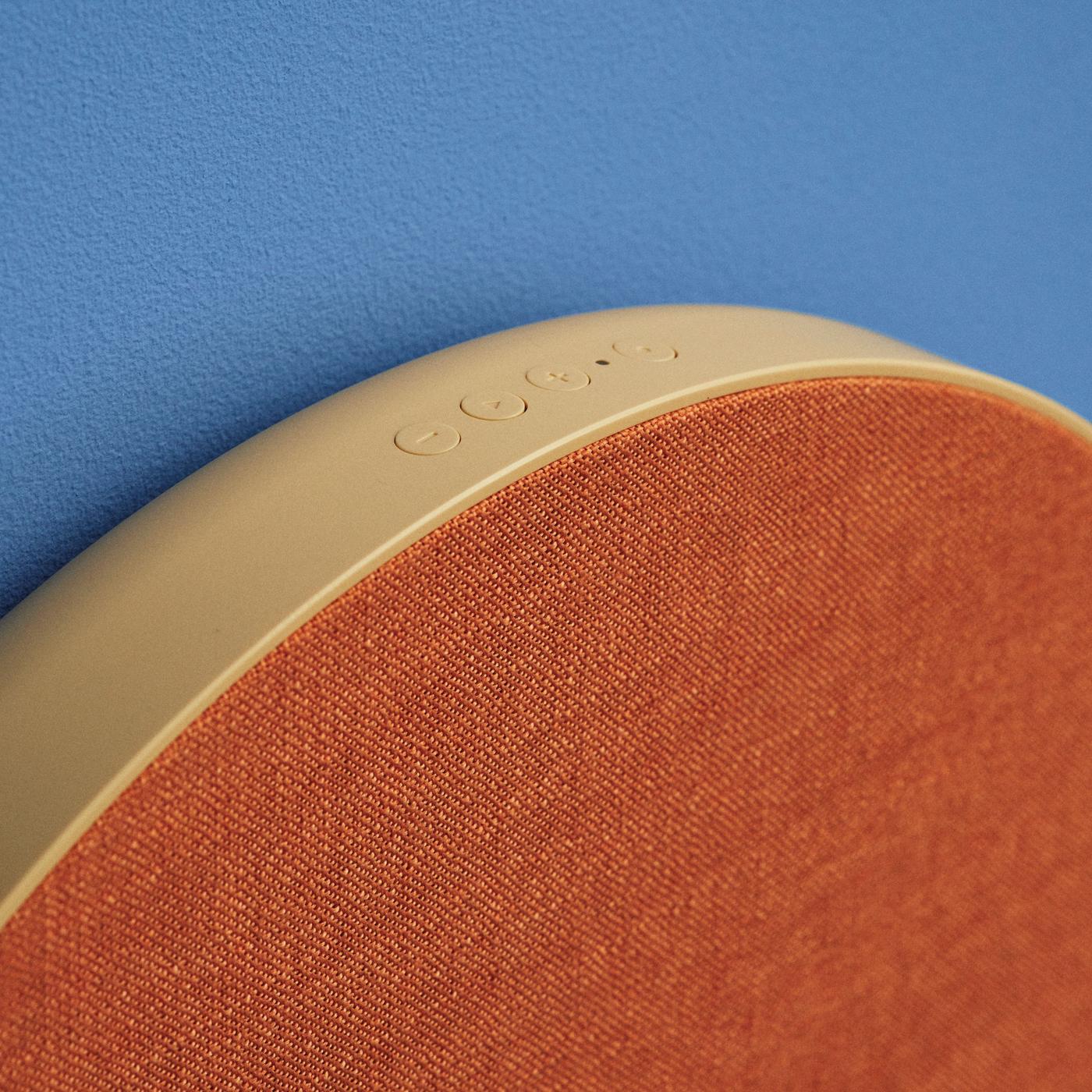 Top and side view of a large round SOLSKYDD speaker with orange textured surface and yellow-beige sides, showing control buttons on top, set against a blue background.