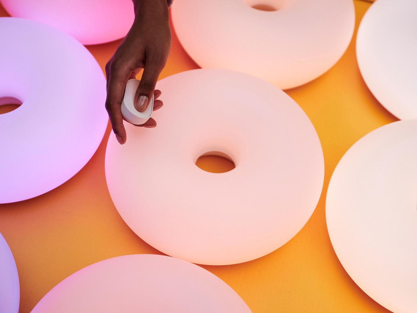 A hand holding a small white remote above glowing donut-shaped lamps in pastel tones on a yellow surface.
