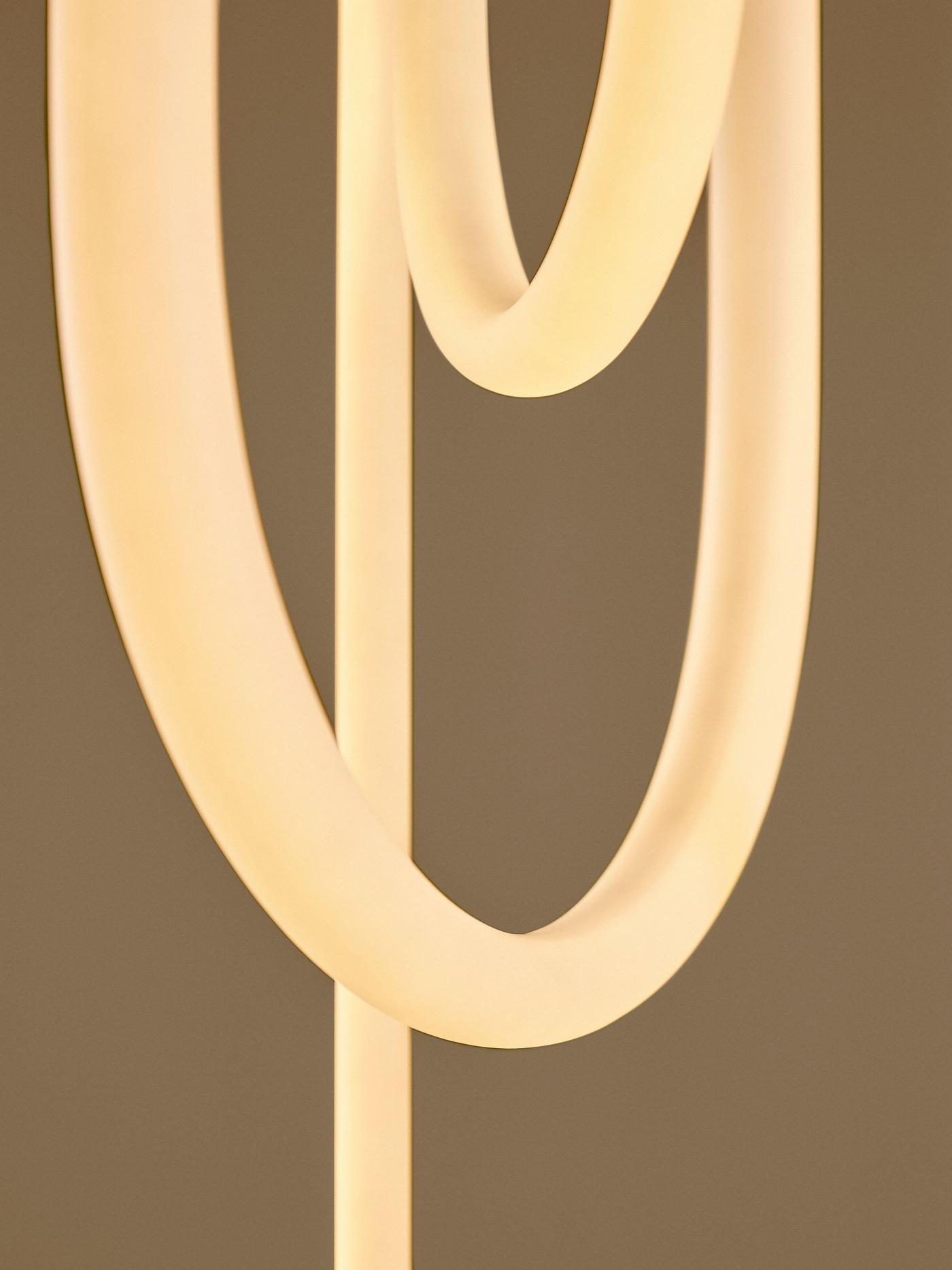 Close-up of curved tubular pendant lamp with soft warm light, in multiple loops against a beige background.