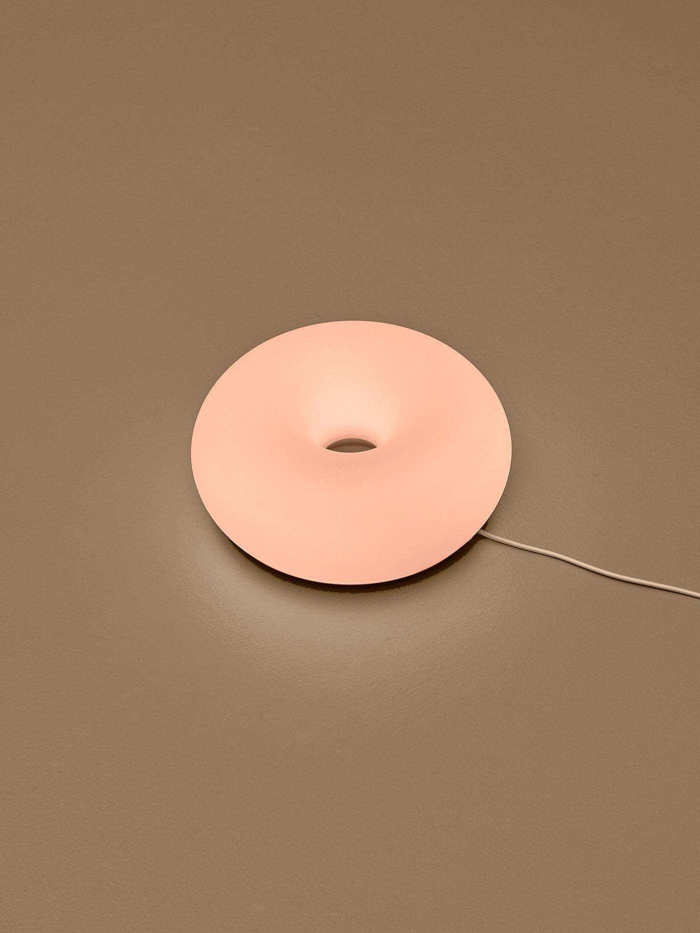 Glowing peach-pink donut-shaped lamp mounted on a coloured wall, with its power cord extending to the side.