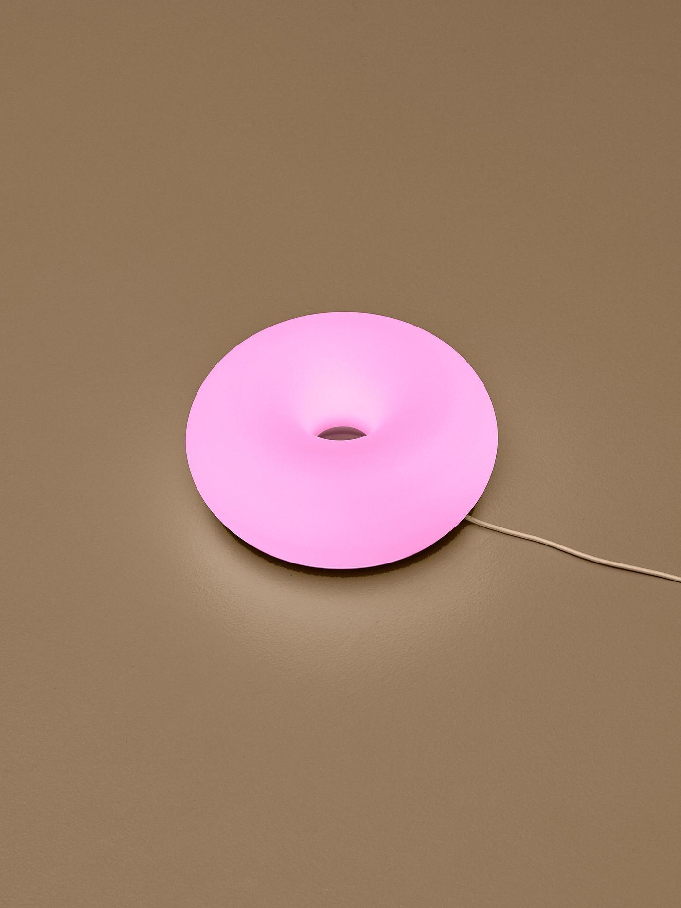 Glowing pink donut-shaped lamp mounted on a coloured wall, with its power cord extending to the side.