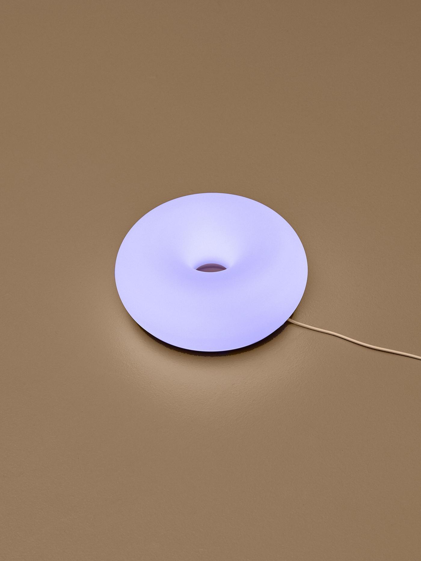 A glowing violet-blue donut-shaped lamp mounted on a coloured wall, with its power cord extending to the side.