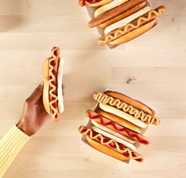 A hand holding a hot dog with ketchup and mustard in a bread and more hot dogs are sitting on a table below.