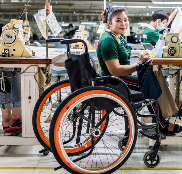 A woman in a green shirt at a sewing machine with a wheel chair in front
