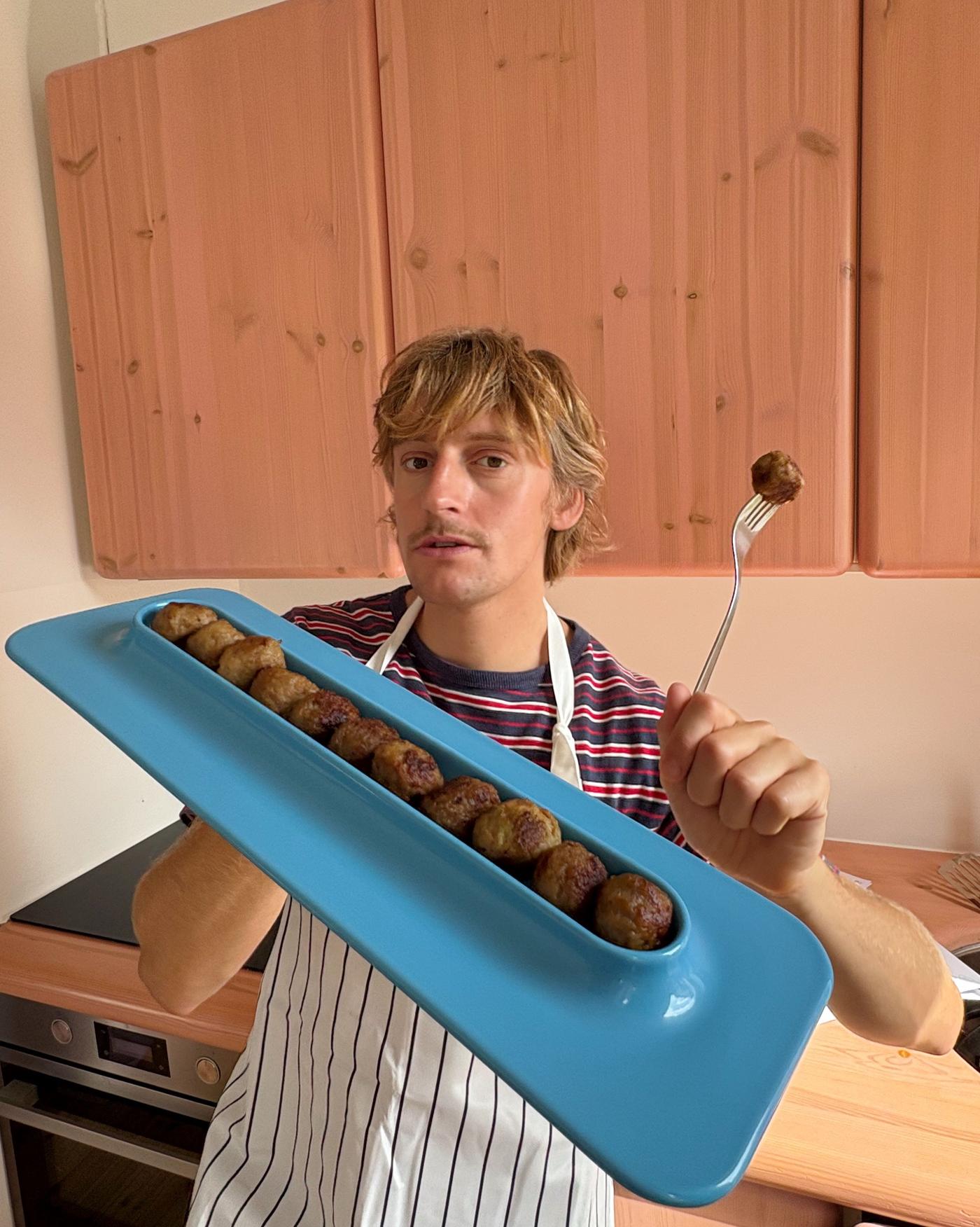 Designer Gustaf Westman in an apron, holding an oblong blue plate like a waiter, with a fork spearing a meatball.