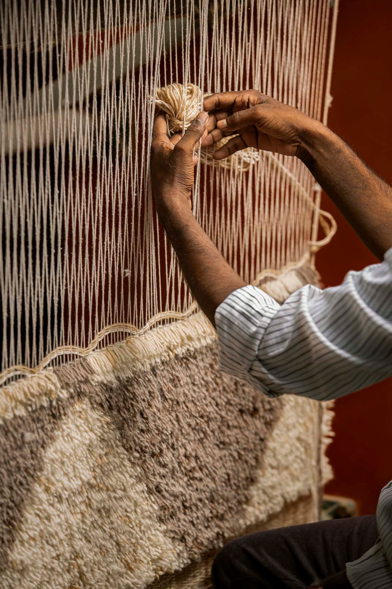 A pair of hands pulls a ball of wool through the strings of the loom to create a hand-knotted patterned rug.