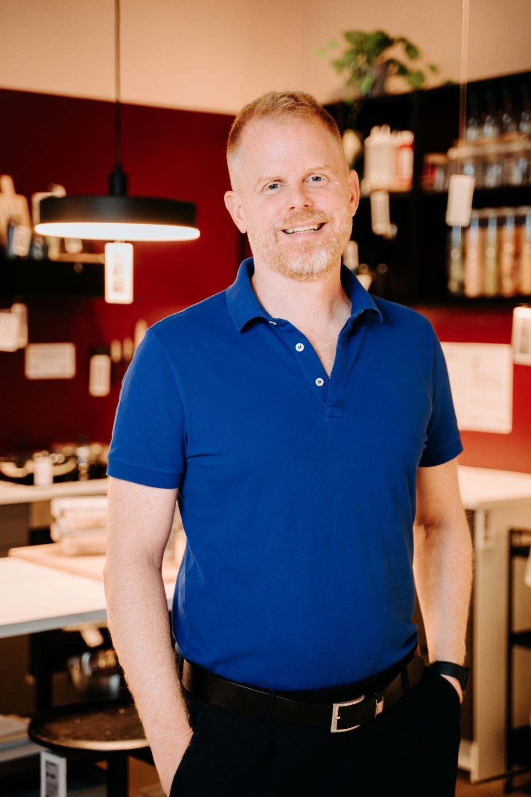 Portrait of Jon Abrahamsson Ring, CEO of Inter IKEA Group, standing and smiling in front of a home furnishing background. He is wearing a blue T-shirt and black pants.