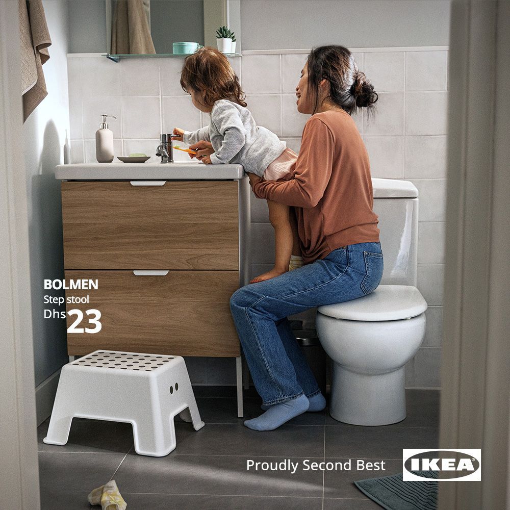 In a bathroom, a mother is seated on a toilet with a small child standing on her lap to reach the tap of the nearby basin. An unused step stool is on the floor nearby.
