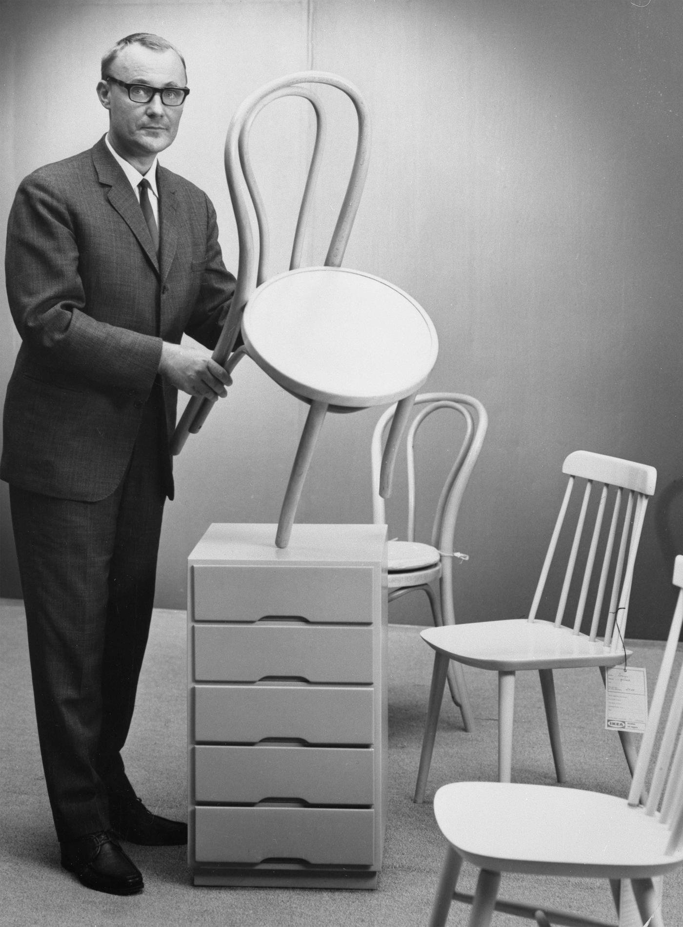 A black-and-white image of Ingvar Kamprad holding a wooden chair, surrounded by chairs and a chest of drawers.